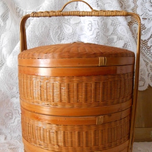 May include: A three-tiered cylindrical woven basket with a light brown finish. The basket features a flat lid, a sturdy arched handle extending from the base to the top, and horizontal bands separating each stackable section with woven texture.