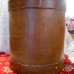 May include: A tall, cylindrical wooden box with a warm brown finish. The box has a lid and a base, and it sits on a red floral patterned fabric. The box appears to be vintage or antique.