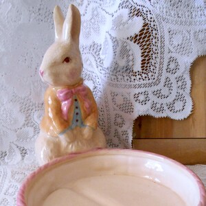 May include: A ceramic dish featuring a sculpted rabbit figure wearing a tan jacket with a pink bow tie. The round, shallow bowl has a textured pink exterior resembling a woven basket and a smooth, cream-colored interior divided into two sections.