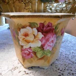 May include: A hexagonal ceramic planter with a hand-painted floral design featuring roses in shades of white, pink, and burgundy. The planter has a cream-colored background and a gold trim. The planter is sitting on a white lace tablecloth.