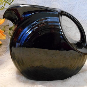 May include: A glossy black pitcher with a distinctive handle and spout. The pitcher has a rounded body and a smooth, reflective surface. A vibrant orange and yellow flower arrangement sits nearby.