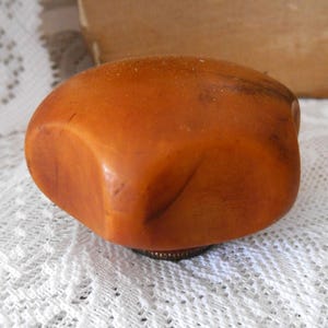 May include: A smooth, rounded, amber-colored object with a polished surface and subtle dark inclusions. The piece features a slightly irregular, organic shape and is attached to a small, circular, metallic base at the bottom.