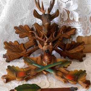 May include: A carved wooden wall decoration featuring two deer heads mounted on oak leaf plaques. The upper deer is dark brown, while the lower deer is lighter with painted green and orange rifles crossed beneath it. A separate leaf-shaped pendulum hangs below.