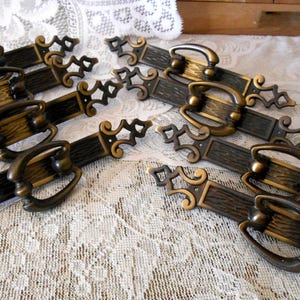 May include: A set of six elongated metal drawer pulls featuring a dark bronze finish with textured centers and ornate, scrolled ends. Each pull includes a central swinging bail handle attached to a decorative backplate with pointed, geometric details.