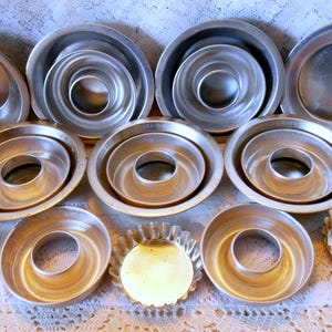 May include: A collection of various sized, silver-colored metal baking pans. The assortment includes round, oval, and fluted pans, including bundt cake pans and tart pans. The pans are arranged on a white lace surface.