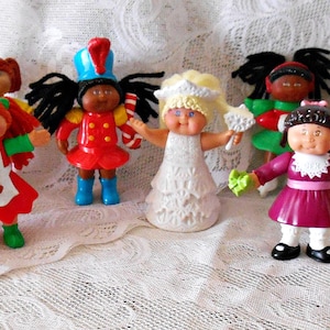 May include: A collection of vintage doll figurines, each with unique outfits and hairstyles. The dolls are in various colors, including pink, red, and yellow, and are arranged on a white lace surface. The dolls have outstretched arms.