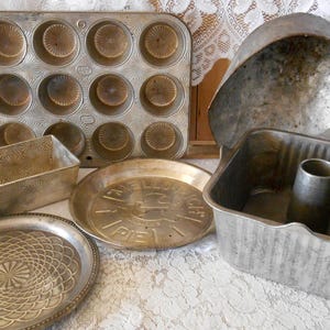 May include: A collection of vintage metal bakeware including a twelve-cup muffin tin, a rectangular loaf pan, two round shallow pans with embossed patterns and text, and a large square cake pan with a central tube and a matching domed lid.