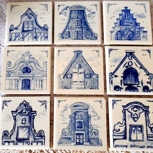 May include: Nine square ceramic tiles with blue and white illustrations of Dutch houses. Each tile features a different architectural design, with detailed drawings of windows, doors, and rooflines. The tiles are arranged on a white lace surface.