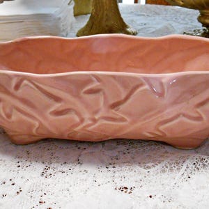 May include: A vintage, oblong, peach-colored ceramic planter. The planter has a wavy rim and is supported by three small feet. The exterior features a raised leaf design. This decorative piece is suitable for indoor or outdoor use.