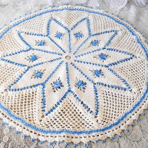 May include: A round, off-white crocheted doily with a central starburst design in light blue. The starburst is outlined and detailed with blue, and the edge is finished with a blue scalloped border. The doily is displayed on a white lace surface.