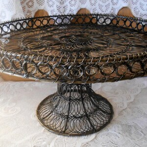 May include: A dark-colored metal pedestal stand featuring a circular tray with a raised rim and a wide, flared base. The entire structure is constructed from interconnected wire coils, creating a repeating circular pattern throughout the surface.