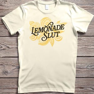 May include: Cream-colored t-shirt featuring the text "LEMONADE SLUT" in a stylized font, accompanied by illustrations of lemons and leaves. The design is in shades of yellow and brown, creating a vintage aesthetic. The shirt has short sleeves and a crew neck.