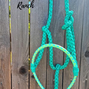 May include: A teal braided rope horse halter featuring a circular noseband with a yellow and teal checkered pattern. The halter includes knotted sections and a white tassel at the bottom. The text Rockin' Heart Ranch is visible in the upper left.