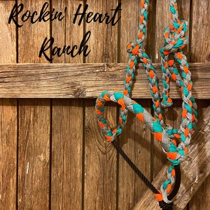 May include: A braided rope halter in teal, orange, and gray with a black tassel hanging from the bottom. The halter is hanging on a wooden fence with the text "Rockin' Heart Ranch" above it.