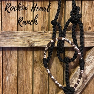 May include: Black and white braided rope halter with a cowhide accent hanging on a wooden fence. The sign above reads "Rockin' Heart Ranch".