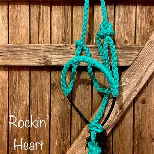 May include: A turquoise braided rope halter with a black rope tied to it. The halter is hanging on a wooden fence. The text "Rockin' Heart Ranch" is written below the halter.