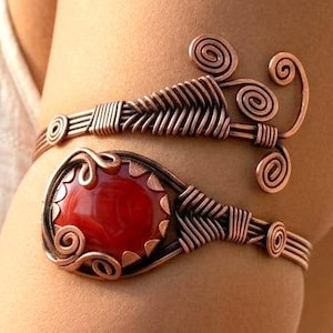 May include: Copper arm cuff featuring a large, oval, red gemstone. The cuff has intricate wirework with spiral and woven designs. The jewelry is designed to be worn on the upper arm.