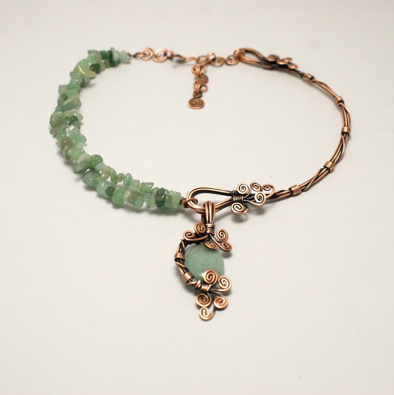 Peut inclure: Un collier en fil de cuivre avec un pendentif en pierre verte. Le collier est fait d'un fil de cuivre torsad&eacute; et a une rang&eacute;e de pierres vertes le long du dos. Le pendentif est une pierre verte envelopp&eacute;e de fil de cuivre avec un motif en fil de cuivre.