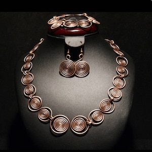 May include: A copper necklace, bracelet, and earrings set with a spiral design. The jewelry is displayed on a black velvet stand.