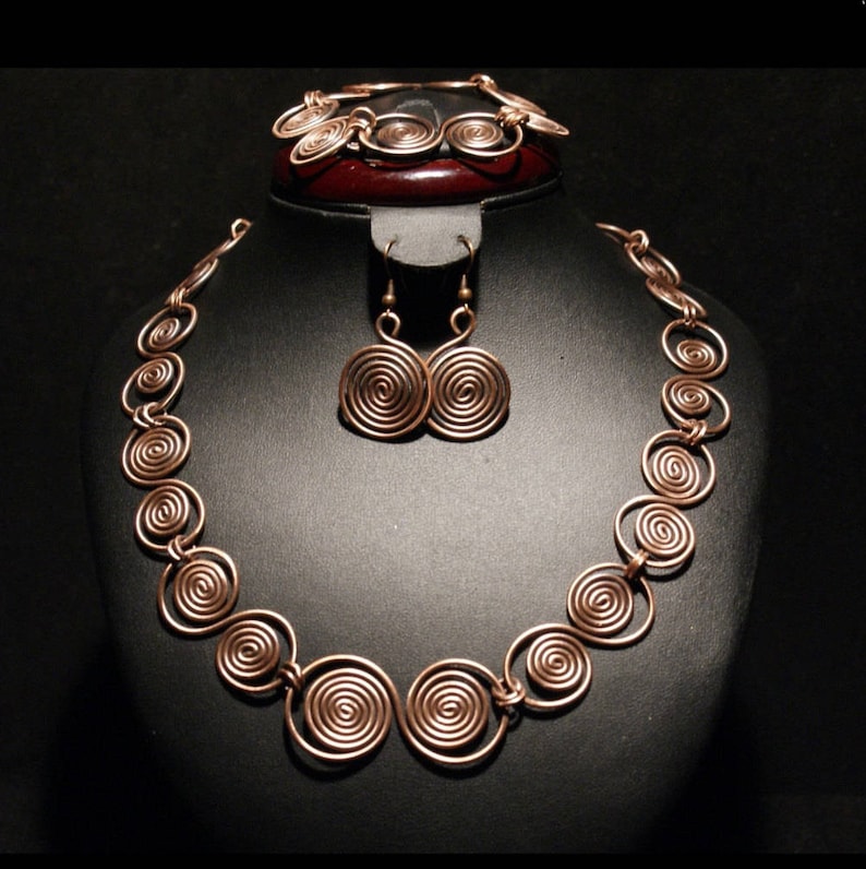 May include: A copper necklace, bracelet, and earrings set with a spiral design. The jewelry is displayed on a black velvet stand.