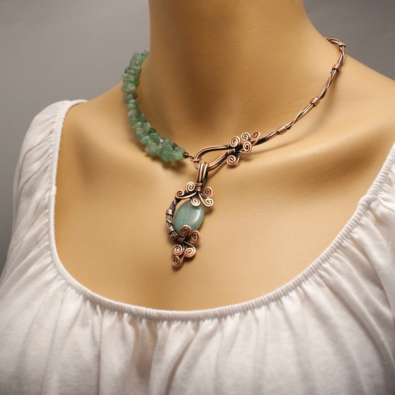 Peut inclure: Un collier en fil de cuivre avec un pendentif en pierre verte et des perles en pierre verte. Le pendentif est fait de fil de cuivre et a un design en spirale. Le collier est sur un fond blanc.