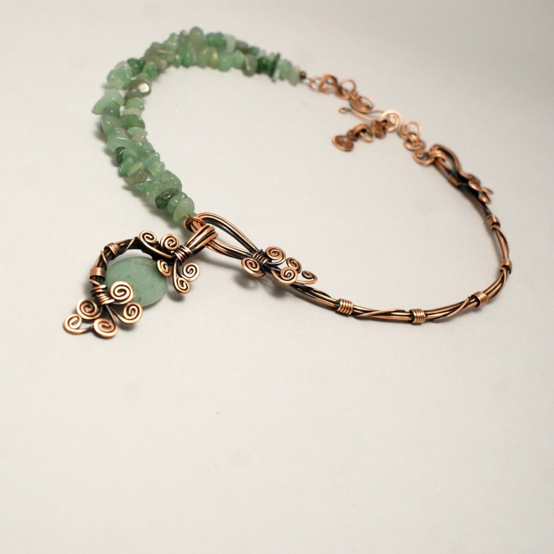 Peut inclure: Un collier en fil de cuivre avec un pendentif en pierre verte. Le pendentif est en forme de branche d'arbre avec des d&eacute;tails en fil de fer boucl&eacute;. Le collier est fait avec une double rang&eacute;e de pierres vertes.