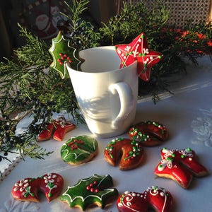 May include: A white mug with a red and white star-shaped biscuit resting on the rim. The mug is surrounded by a variety of Christmas-themed biscuits, including holly leaves, stars, and reindeer. The biscuits are decorated with green, red, and white icing.