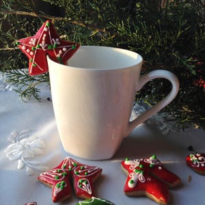 May include: A white ceramic mug with a red and green star-shaped gingerbread cookie resting on the rim. The cookie is decorated with white icing and green and red sprinkles. Other gingerbread cookies are visible in the background.