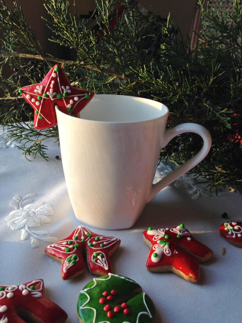 May include: A white ceramic mug with a red and green star-shaped gingerbread cookie resting on the rim. The cookie is decorated with white icing and green and red sprinkles. Other gingerbread cookies are visible in the background.