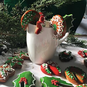 May include: A white mug filled with gingerbread cookies decorated with green holly leaves, red berries, and white icing. The cookies are arranged around the mug on a white tablecloth.