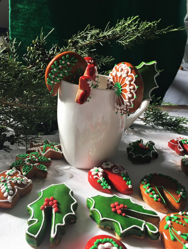 May include: A white mug filled with gingerbread cookies decorated with green holly leaves, red berries, and white icing. The cookies are arranged around the mug on a white tablecloth.