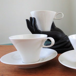 Può includere: Tazzine e piattini in porcellana bianca. Le tazzine hanno una forma conica con un manico curvo. I piattini sono rotondi e piatti. Le tazzine e i piattini sono disposti su una superficie di legno.