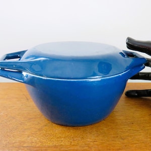 Pode incluir: Uma caçarola azul cobalto com tampa e alças integradas. A travessa de cerâmica está sobre uma superfície de madeira. Uma escultura preta de uma mão está ao lado da caçarola.