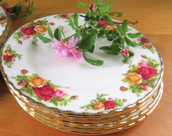 May include: A stack of white porcelain plates with a gold rim and a floral rose pattern in red, yellow, and green. The plates are arranged on a wooden surface, with a sprig of pink flowers and green leaves.