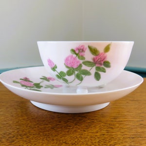 May include: A white teacup and saucer set with a pink floral design. The teacup is round and sits on a matching saucer. The floral design features pink flowers and green leaves. The set is on a wooden surface.