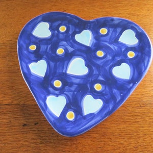 Pode incluir: Um prato de cerâmica em forma de coração com fundo azul escuro. O prato apresenta corações azuis claros e pontos amarelos. O prato está sobre uma superfície de madeira.