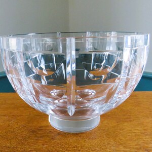 Puede incluir: Un cuenco de cristal transparente y facetado con una forma ancha y redondeada. El cuenco presenta un patrón geométrico de cuadrados y líneas, y se asienta sobre una pequeña base circular. El cuenco se exhibe sobre una superficie de madera.