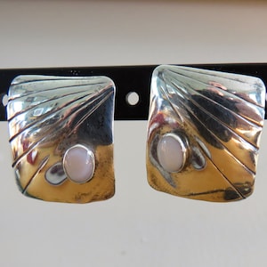 Peut inclure: Une paire de boucles d'oreilles carrées argentées et dorées. Chaque boucle d'oreille présente une pierre ovale rose clair au centre. La partie argentée a un motif texturé en forme d'éventail. Les boucles d'oreilles sont exposées sur un présentoir noir.