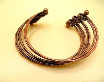 Peut inclure: Bracelet manchette en cuivre avec plusieurs brins de fil. Le bracelet présente un motif torsadé à une extrémité et des boucles à l'autre, avec de petits accents de boules. Le métal brun rougeâtre chaud est présenté sur un fond jaune clair.