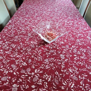 May include: A rectangular burgundy tablecloth with a white floral pattern, including leaves, berries, and pinecones. A clear, square glass bowl filled with food sits on the tablecloth. The table is set for a meal.