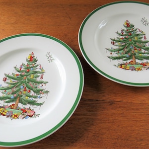Op de afbeelding: Twee witte keramische borden met een groene rand. Elk bord heeft een gedetailleerde illustratie van een versierde kerstboom met ornamenten en cadeaus. De borden staan op een houten oppervlak.
