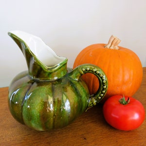 Puede incluir: Una jarra de cerámica verde con forma de calabaza, interior blanco y asa con cuentas. Una calabaza naranja y un tomate rojo están junto a la jarra. Los objetos están sobre una superficie de madera.
