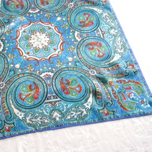 Puede incluir: Un textil cuadrado con un estampado de cachemira en tonos azules, turquesas y blancos. El diseño presenta grandes espirales con detalles rojos y verdes. Los bordes están acabados con un fino borde azul. La tela parece suave y ligera.