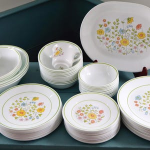 Könnte beinhalten: Ein großes Set aus weißem Geschirr mit einem Blumenmuster in Gelb, Orange, Blau und Grün sowie dünnen grünen Rändern. Das Set besteht aus Speisetellern, Salattellern, Schüsseln, Tassen, Untertassen und einer großen ovalen Servierplatte.