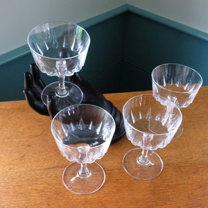 Peut inclure: Quatre coupes à champagne transparentes avec un motif en étoile sur le bol. Les verres sont posés sur une surface en bois, l'un d'eux étant tenu par une sculpture de main noire. Design classique et élégant.