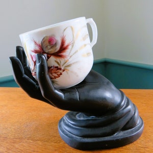 Peut inclure: Une tasse blanche avec un motif floral rouge, marron et doré est tenue par une sculpture noire en forme de main. La tasse a une anse incurvée et est inclinée, mettant en valeur les détails. La sculpture repose sur une surface en bois.