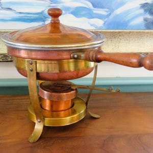 Może przedstawiać: Miedziany i mosiężny podgrzewacz do potraw z drewnianym uchwytem i pokrywką. Naczynie ma okrągłą podstawę i mały palnik poniżej. Pokrywka ma drewniane pokrętło. Podgrzewacz do potraw jest przeznaczony do utrzymywania ciepła potraw.