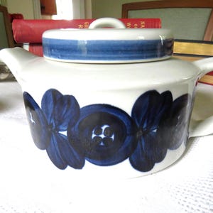 Könnte beinhalten: Eine weiße Keramik-Teekanne mit einem dunkelblauen Blumenmuster um den Körper und einem passenden Deckel mit blauem Band. Die Teekanne hat einen geschwungenen Griff auf der rechten Seite und einen kurzen Ausguss auf der linken Seite mit glatter Oberfläche.