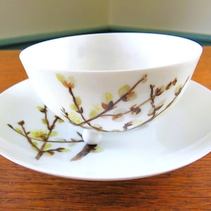 May include: A white teacup and saucer set. The cup and saucer feature a delicate floral design in yellow and brown. The cup rests on a small, white, spherical base, and the saucer is round. A classic tea set.