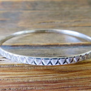 May include: A silver bangle bracelet with a geometric pattern. The bracelet is round and has a series of engraved triangles and lines around the band. The bracelet is displayed on a wooden surface.